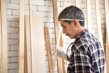 Senior asian old man carpenter wearing glasses and gloves protection build diy furniture select wood plank in workshop