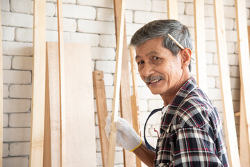 Senior asian old man carpenter wearing glasses and gloves protection build diy furniture select wood plank in workshop