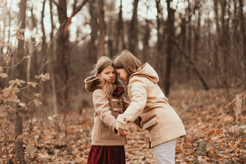 Caucasian children sisters walk together in the park in the forest, girlfriends children hold hands and hug for a walk, girly secrets, autumn walks in warm coats
