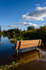 Wooden bench on the flooded shore of Larson Lake, reflections of blue sky and clouds
