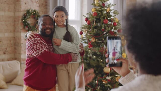 Cheerful Afro-American man and his pretty daughter standing by Christmas tree at home, embracing and smiling while mother taking picture of them with smartphone