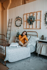 Happy woman in orange sweater on the old fashioned bed with many pillows. Rustic bedroom with christmas decorations. New year eve in the village. Woman in village house.
