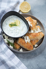 Greek baked mini filo rolls filled with feta and served with tzatziki dip, vertical shot on a light-blue stone background