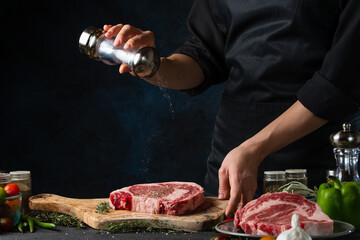 Close-up view of chef salts raw steak on wooden chopped board. Backstage of preparing grilled pork meat at restaurant kitchen on dark blue background. Frozen motion. Cooking process.