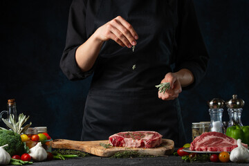 Chef pours rosemary on raw steak on wooden chopped board. Backstage of preparing grilled pork meat at restaurant kitchen on dark blue background. Frozen motion. Cooking process.