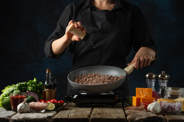 Close-up view of professional chef in black uniform salts meat filling for cooking tacos on dark blue background. Traditional mexican cuisine. Concept of tasty street food. Frozen motion.