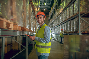 Male warehouse worker in orange helmet standing near containers and holding a tablet