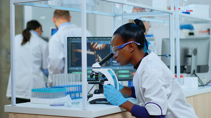 Black biochemist woman sitting in modern equipped lab analysing blood tests. Multiethnic team...