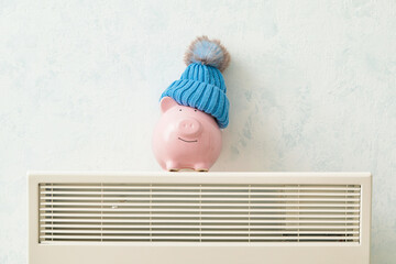 Piggy bank and hat on radiator. Concept of heating season