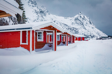 Landscape photography. Traditional Norwegian red wooden houses under the fresh snow. Gloomy winter scene of Lofoten islands on the shore of Kongsjordpollen fjord, Vestvagoy, Norway.