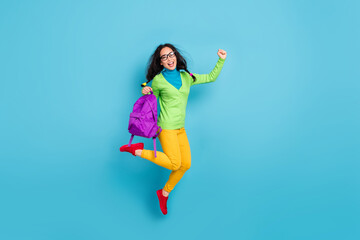 Full size photo of cheery schoolgirl hold bag raise fist wear green shirt yellow trousers isolated on blue color background