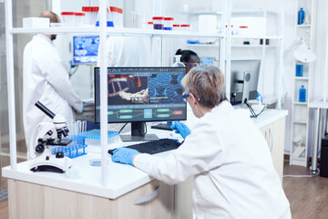Senior scientist in pharmaceuticals laboratory doing genetic research wearing lab coat with team in the background. Male chemist engineering vaccine working on computer. - obrazy, fototapety, plakaty
