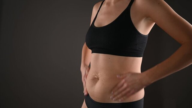 The young woman shows changes in her figure and does a hand massage, flabby saggy belly. Concept of body care, proper nutrition, excess weight, women's health. Woman in underwear on grey background. 