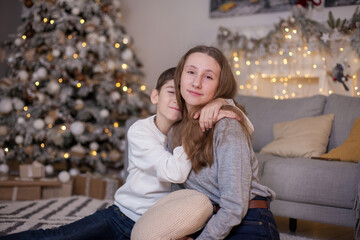 happy brother and sister sitting on floor near sofa and christmas tree with presents and looking at camera. Boy and girl at Christmas time. Merry Christmas and Happy New Year