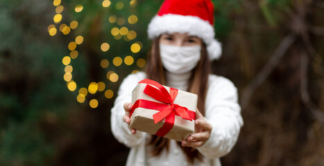 Christmas mask congratulations. Portrait woman wearing santa hat and white sweater in medical mask, giving gift present box with red ribbon, christmas tree bokeh on background