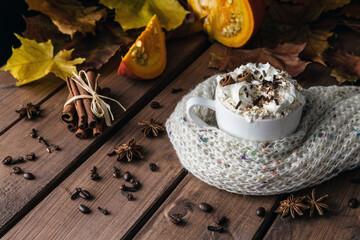 Cup of coffee with cinnamon sticks, whipped cream and chocolate sprinkles. Mug covered with knitted winter scarf. Cozy autumn composition of hot beverage on wooden table.