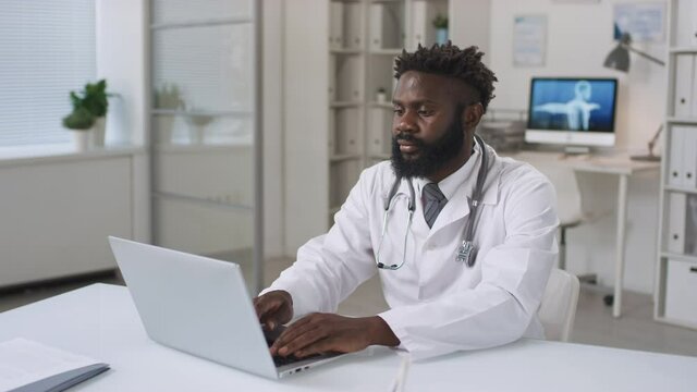 Professional African American male medical specialist sitting at desk in doctors office working on medical records using laptop