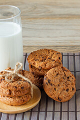 A stack of chocolate chips cookies on baking paper, with a glass of milk on a light wooden background