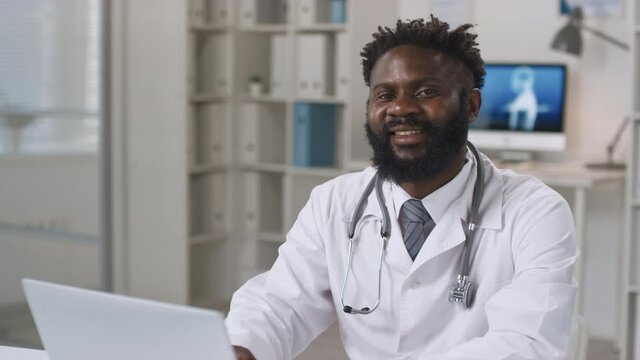 Portrait shot of young adult African American male doctor wearing white coat and stethoscope around neck sitting at desk working with data on laptop then smiling at camera
