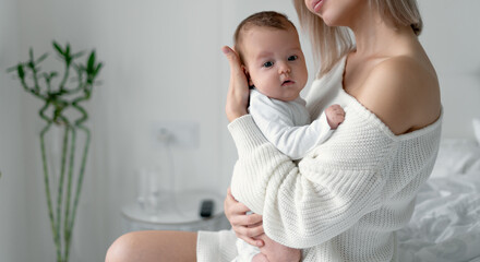 A young mother holds a newborn baby in her arms and presses her head to her chest. Woman and child in the bedroom on a white background. Motherhood and childhood concept.