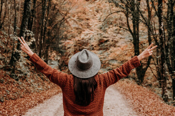 Female with open arms wearing a brown jacket with a fedora hat in a forest in autumn - obrazy, fototapety, plakaty