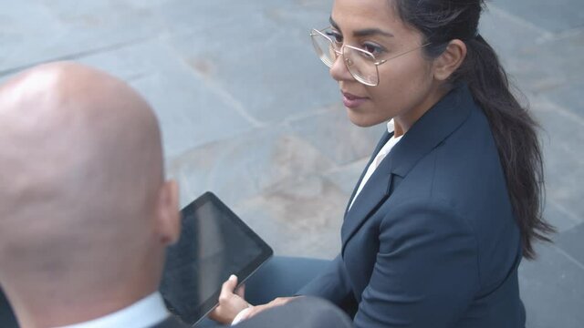 Focused pretty Latin businesswoman listening to male colleague, holding tablet while sitting outside. High angle. Business meeting and communication concept