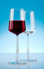 Two wineglasses on reflective surface. One filled with red wine and on empty. Studio photo isolated on blue background.