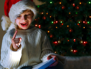 Cute boy in red santa hat and white sweater writing letter to Santa on the background of a Christmas tree decorated with lights with place for text. The kid got the idea. Copyspace.