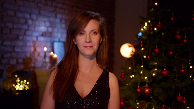 Happy woman wishing Merry Christmas and Happy New Year. Portrait of woman in elegant black dress at home with Christmas tree.