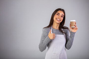 Young waitress of cafe in apron giving you cup of hot coffee while standing in front of camera in isolation. A waitress holding and serving a paper cup of hot coffee