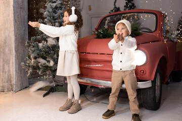 Children playing with snow near red car and Christmas tree and lights on background. Merry Christmas and Happy Holidays
