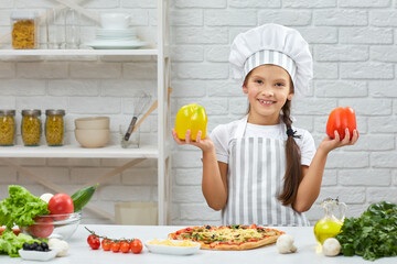 the child holding yellow and red bell peppers. little girl in chef hat and an apron cooking pizza in the kitchen.