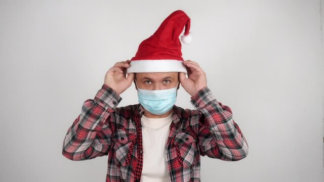 A man puts on a santa claus hat and raises his hands up. Happy man in medical mask celebrating Christmas. 