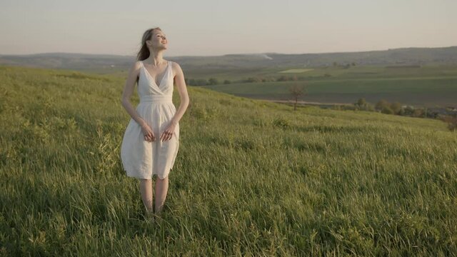 shy girl in a white dress is thrilled by the air currents on the meadow