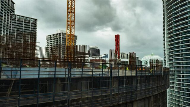 Time lapse construction workers in Miami build a luxury condo and pour concrete foundation and pillars with help from cranes as clouds roll past on an overcast afternoon featuring a dark ominous sky 