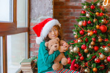 little Asian girl in pajamas sitting on the window near the Christmas tree with Teddy bear Christmas, new year