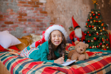 little Asian girl in pajamas near the Christmas tree writes a letter to Santa Claus Christmas, new year