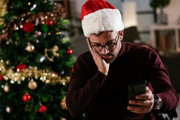 Young sad man in wheelchair sitting near Christmas tree..