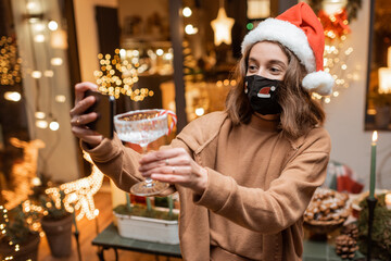 Young woman in facial mask celebrating alone New Year holidays at home, having a video call on the phone with friends. Concept of quarantine and self-isolation during the epidemic on holidays