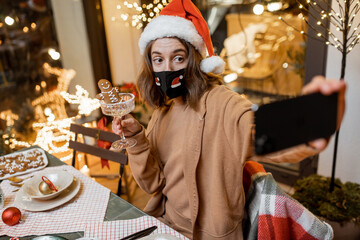 Young woman in facial mask celebrating alone New Year holidays at home, having a video call on the phone with friends. Concept of quarantine and self-isolation during the epidemic on holidays
