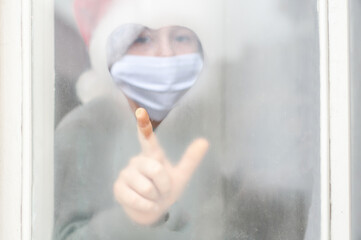 Child boy girl a santa claus hat, protective mask draws a heart on the foggy misted window with his...