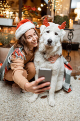 Portrait of a woman in christmas hat with her cute dog celebrating a New Year holidays on the beautifully decorated terrace at home, making selfie photo together