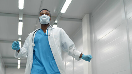 Fototapeta premium Young afro doctor man wearing hospital coat, safety mask and gloves dancing in corridor
