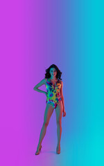 Smiling. Beautiful seductive girl in fashionable bright colourful swimsuit on gradient pink-blue neon background. Full-length portrait. Copyspace for ad. Summer, fashion, emotions concept.