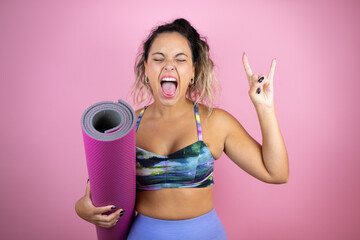 Young beautiful woman wearing sportswear and holding a splinter over isolated pink background shouting with crazy expression doing rock symbol with hands up