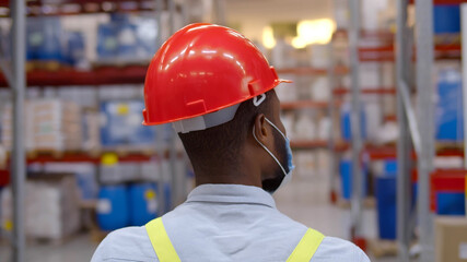 Black male worker wearing protective face mask and hardhat walking in factory warehouse.