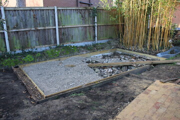 Close up of shed base construction of wood edging prepared hardcore mortar concrete flint brick compressed surface , a gravel surround french drain & brown earth soil & garden fencing background