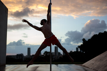 Muscular male pole dancer, holding a pose on a pole set outdoors. Silhouette shot with Tel Aviv buildings in the far background.