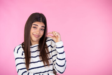 Size matters. Pleased brunette young woman demonstrates very tiny object, smiles positively, wears casual sweater, poses against pink background, shapes small thing. Body language concept - obrazy, fototapety, plakaty