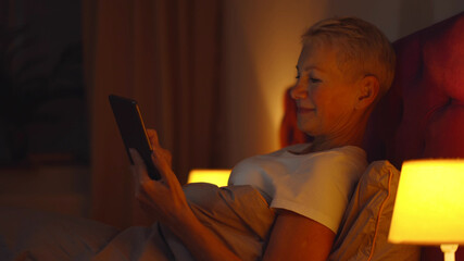 Joyful beautiful senior woman using smartphone sitting on bed at home in evening